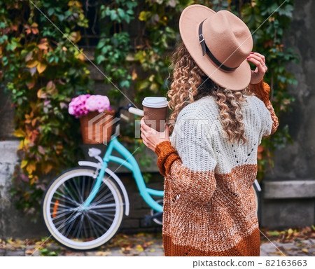 Romantic atmosphere in style of Provence. Rear view of woman enjoying coffee and adjusting her hat on head. On blurred backdrop wall with green ivy plants and parked to it city retro bicycle. 82163663