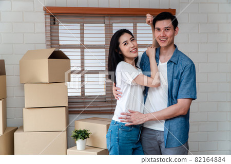 Smiling young Asian happy couple hugging boyfriend with cardboard boxes at moving day in their new home after buying real estate. Concept of starting a new life for a newly married couple. 82164884