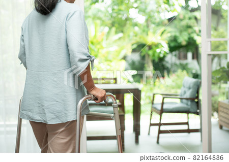 A sad old elderly woman uses Walker for standing in front of windows and looking outside and feeling lonely. Concept of depression caused by illness and Elderly caregivers A sad old elderly woman uses Walker for standing in front of windows and looking outside and feeling lonely. Concept of depression caused by illness and Elderly caregivers 82164886