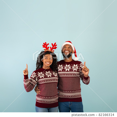 Holiday offer. Happy black spouses in identical Christmas sweaters pointing up at copy space, blue background Holiday offer. Happy black spouses in identical Christmas sweaters pointing up at copy space, blue background 82166314