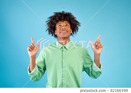 Positive african american guy pointing fingers up above his head, posing over blue background Positive african american guy pointing fingers up above his head, posing over blue background 82166399