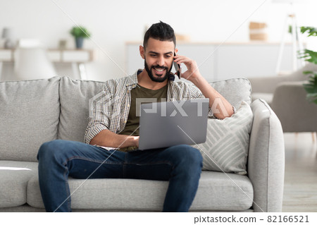 Happy young Arab man using laptop for video conference, calling on smartphone, working online from home Happy young Arab man using laptop for video conference, calling on smartphone, working online from home 82166521