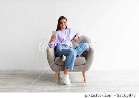 Smiling millennial woman chatting online on smartphone, sitting in comfy armchair on white studio background Smiling millennial woman chatting online on smartphone, sitting in comfy armchair on white studio background 82166975