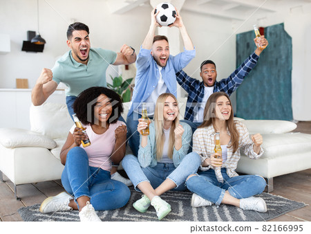 Emotional diverse friends watching soccer on TV, drinking beer, holding ball and shouting, celebrating victory at home 82166995