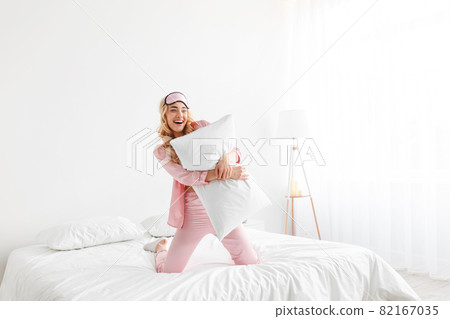 Funny happy millennial pretty caucasian blonde female in pink pajama, sleep mask, hug pillow, have fun on comfort bed 82167035