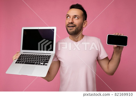 Handsome man holding laptop computer and mobile phone looking up in t-shirt on isolated pink 82169910