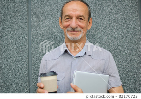 Male portrait of older coworker with silver laptop and cup of coffee waiting for a meeting near dark grey wall in the morning, remote job, coffee break Male portrait of older coworker with silver laptop and cup of coffee waiting for a meeting near dark grey wall in the morning, remote job, coffee break 82170722