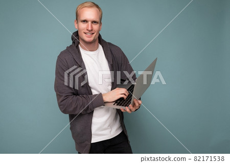 Photo of handsome blond man holding and using computer laptop looking at camera isolated over blue Photo of handsome blond man holding and using computer laptop looking at camera isolated over blue 82171538