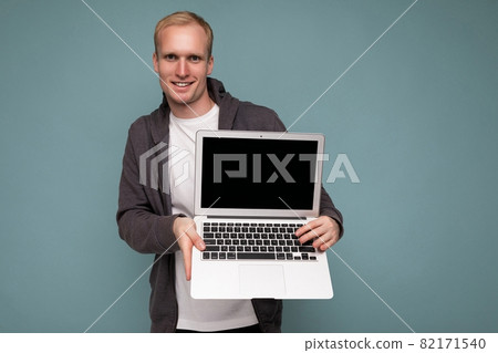 Photo of handsome blond man holding computer laptop looking at camera isolated over blue wall Photo of handsome blond man holding computer laptop looking at camera isolated over blue wall 82171540