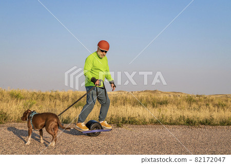 mature senior male is riding one-wheeled electric skateboard  with a dog 82172047