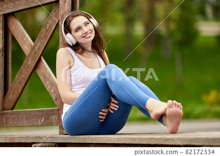Happy shoeless woman in wireless headphones is sitting outdoors. 82172334