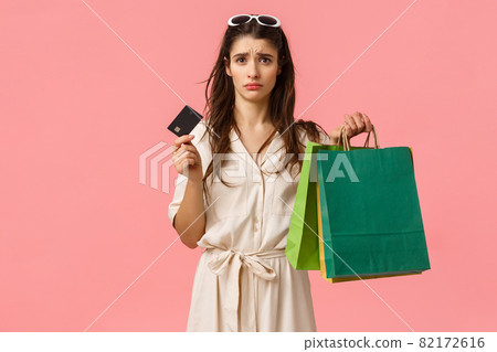 Upset and distressed young brunette female feeling sad spent all money, looking uneasy and concerned at credit card, holding shopping bags, standing pink background gloomy 82172616