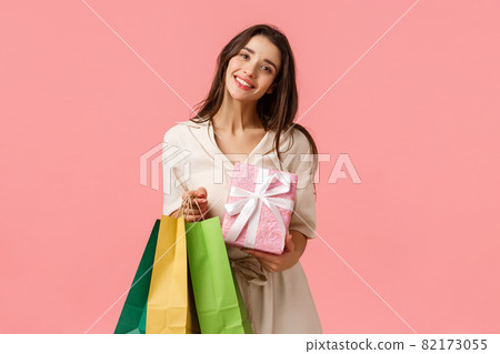 Congratulations, holidays and shopping concept. Charming tender young feminine girl came from store bought lots of gifts, holding wrapped box, standing pink background happy 82173055