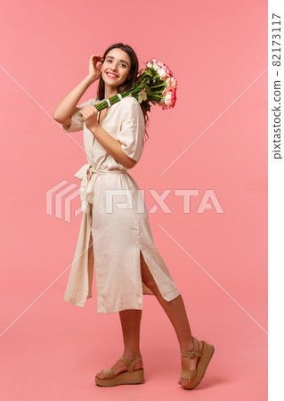 Full-length vertical portrait, dreamy romantic brunette female enjoying date, holding bouquet roses, touching hair tenderly, smiling silly camera, receiving flowers, standing pink background 82173117