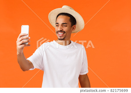 Cheerful happy african-american guy taking selfie on vacation under sun near sandy beautiful beach tropical island, holding smartphone and smiling at mobile camera, standing orange background Cheerful happy african-american guy taking selfie on vacation under sun near sandy beautiful beach tropical island, holding smartphone and smiling at mobile camera, standing orange background 82175699