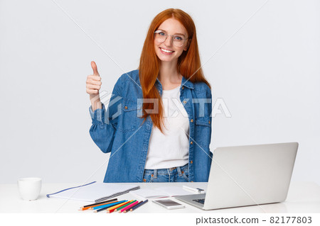 Lovely attractive redhead female in glasses working on project with team member, showing thumb-up checking drawing of friend, standing near colored pencils and laptop, like idea Lovely attractive redhead female in glasses working on project with team member, showing thumb-up checking drawing of friend, standing near colored pencils and laptop, like idea 82177803