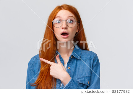 Surprised and wondered, amused attractive redhead woman in glasses, red natural hair, freckles, blue eyes open mouth fascinated and curious, pointing finger left, asking question about banner 82177982