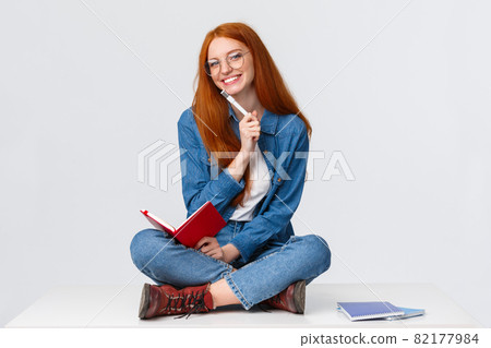 Charismatic, friendly cute redhead girl dreaming, taking notes or prepare homework while sitting on floor with legs crossed, holding notebook and pen, writing over white background 82177984