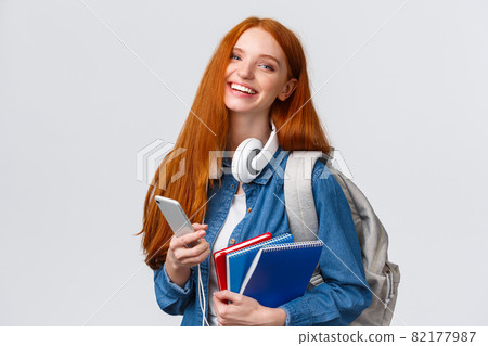 Cheerful, friendly and charismatic redhead woman laughing joyfully, texting friends, chatting with smartphone, enjoying college lifestyle, heading campus with backpack and notebooks 82177987