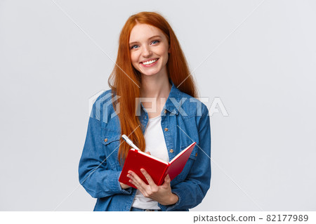 Waist-up portrait lovely redhead teenage girl taking notes during favorite college class, holding pen and red notebook, smiling carefree camera, writing something, make to-do list 82177989