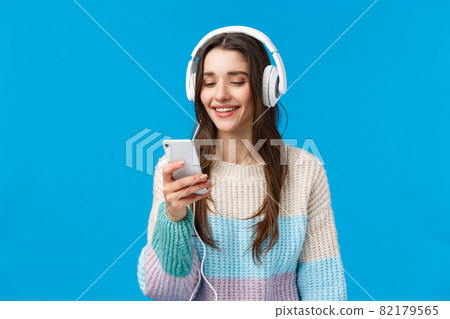 Technology, millennials and lifestyle concept. Carefree cute brunette female student put on headphones, plug-in smartphone picking song and smiling, standing blue background make playlist for study 82179565
