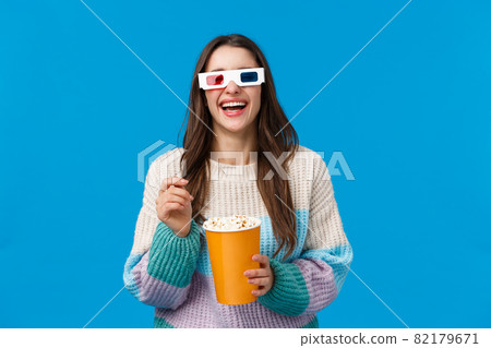 Girl watching comedy in cinema. Carefree happy young brunette woman in winter sweater, enjoying funny movie, laughing out loud eating popcorn, wearing 3d glasses, standing blue background 82179671