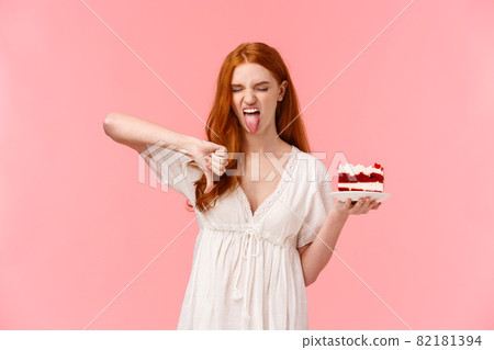 Ignorant, picky and snobbish redhead birthday girl hate b-day cake, holding plate with dessert, showing tongue grimacing and make thumb-down in disapproval, aversion and disgust 82181394