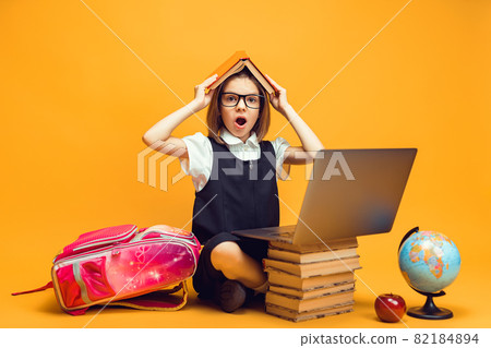 Surprised schoolgirl sits behind a stack of books and laptop holds book on head. Kids education Surprised schoolgirl sits behind a stack of books and laptop holds book on head. Kids education 82184894