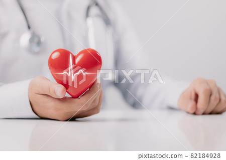 The doctor is holding and showing a red heart with life graph on white background. The doctor is holding and showing a red heart with life graph on white background. 82184928