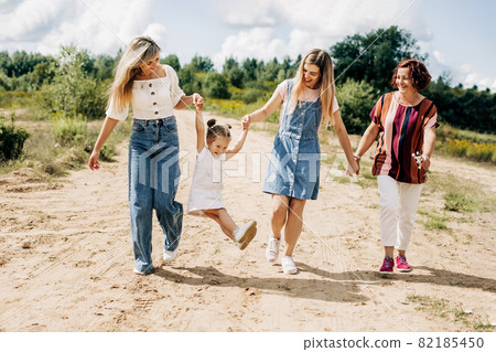 Several generations of women of the same family are walking along a dirt road outside the city, enjoying the time spent together Several generations of women of the same family are walking along a dirt road outside the city, enjoying the time spent together 82185450