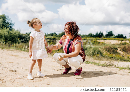Grandmother and granddaughter are walking along a field road outside the city. An elderly woman is playing with a little girl 82185451
