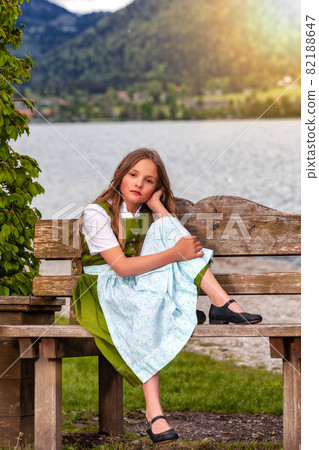 Portrait of girl sitting in dirndl on bench Portrait of girl sitting in dirndl on bench 82188647