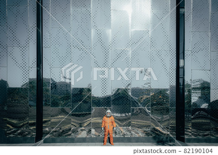 Astronaut wearing orange spacesuit and space helmet standing near the building of a space center outdoors. 82190104