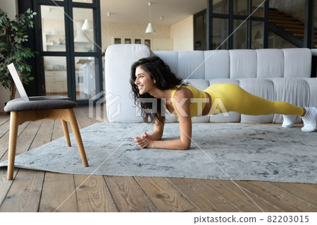 Sporty millennial woman standing in elbow plank in front of laptop, following online sports video at home Sporty millennial woman standing in elbow plank in front of laptop, following online sports video at home 82203015