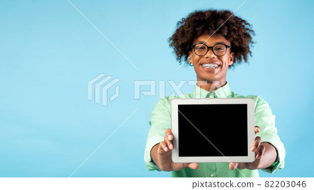 Cheerful black teen guy showing brand new digital tablet with blank screen over blue background, panorama, free space Cheerful black teen guy showing brand new digital tablet with blank screen over blue background, panorama, free space 82203046