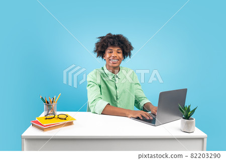 Happy african american teen guy sitting at desk and using laptop, doing homework, studying online, blue background Happy african american teen guy sitting at desk and using laptop, doing homework, studying online, blue background 82203290