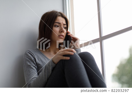 Upset shocked depressed young pretty caucasian female calling by phone, got bad news or breaking up near window 82203297