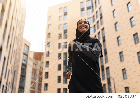 Young Muslim Woman In Modest Sportswear Jogging On City Streets, Low Angle Young Muslim Woman In Modest Sportswear Jogging On City Streets, Low Angle 82203334