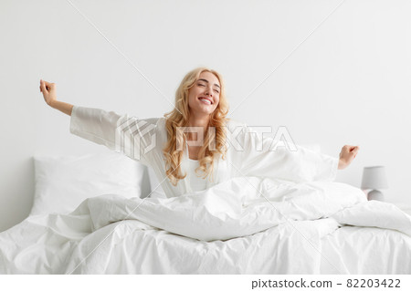 Satisfied millennial attractive energy caucasian blonde female in bathrobe stretching body and enjoy good morning Satisfied millennial attractive energy caucasian blonde female in bathrobe stretching body and enjoy good morning 82203422