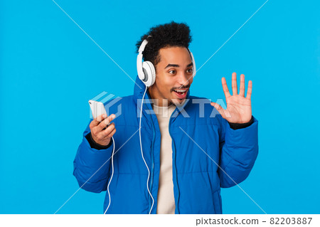 Technology, chatting concept. Stylish attractive young african american male student in padded winter jacket, listening music on way university, hold smartphone headphones, waving hello, hi Technology, chatting concept. Stylish attractive young african american male student in padded winter jacket, listening music on way university, hold smartphone headphones, waving hello, hi 82203887