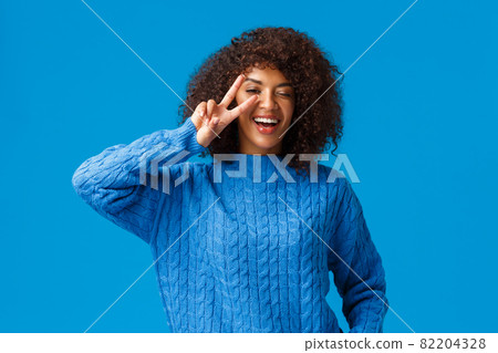 Carefree lovely, charismatic african american woman say cheeze posing delighted and upbeat, showing peace sign over winking eye and smiling, laughing happily, enjoy winter holidays, blue background Carefree lovely, charismatic african american woman say cheeze posing delighted and upbeat, showing peace sign over winking eye and smiling, laughing happily, enjoy winter holidays, blue background 82204328