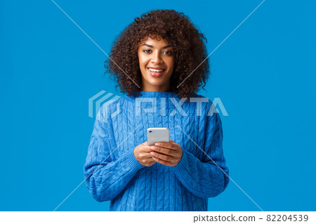 Smartphone addication, gen-z and people concept. Charismatic lovely smiling african-american woman with afro haircut in winter sweater, holding smartphone and looking camera, blue background 82204539