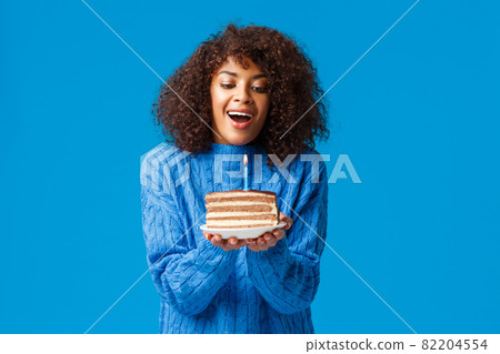 Happy dreamy and hopeful birthday girl making wish. Attractive african american woman with curly haircut, inhale air to blow-out lit candle on tasty b-day cake, standing blue background Happy dreamy and hopeful birthday girl making wish. Attractive african american woman with curly haircut, inhale air to blow-out lit candle on tasty b-day cake, standing blue background 82204554