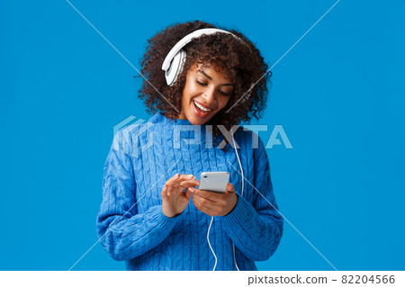 Modern lifestyle, technology and urban concept. Attractive hipster girl african american woman in winter sweater, afro haircut, wearing headphones and messaging using smartphone, smiling 82204566