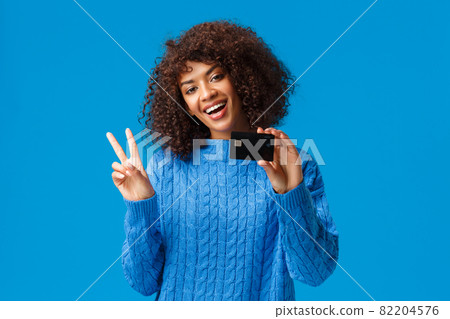 Cheerful cute african-american woman with afro haircut, tilt head show peace sign and holding credit card, got everything for holidays purchase online, standing blue background joyful 82204576