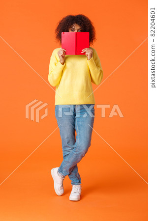 Full-length vertical shot shy and cute african-american modern urban girl with afro haircut, hiding face behind red notebook and peeking camera, writing in diary, standing orange background Full-length vertical shot shy and cute african-american modern urban girl with afro haircut, hiding face behind red notebook and peeking camera, writing in diary, standing orange background 82204601