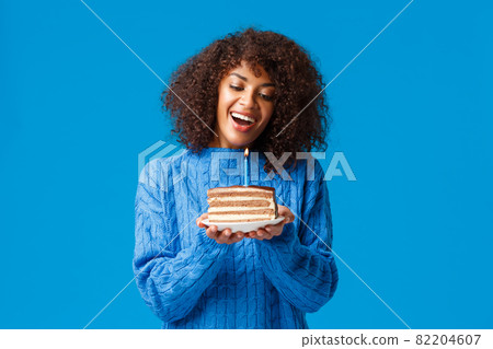 Celebration, holidays and party concept. Dreamy and lovely cute african-american woman with afro haircut, in sweater, tilt head and watching at lit candle on birthday cake, smiling making wish 82204607