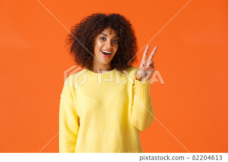 Carefree lovely, silly african-american girl posing for photo over orange background with smile, showing peace sign saying cheese feeling joyful and upbeat, express positive emotions 82204613