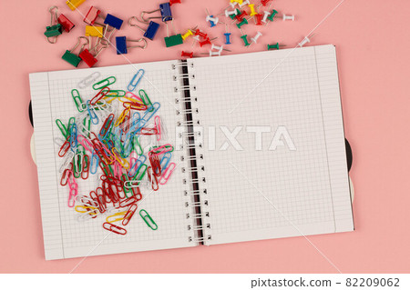 Open school squared notebook and colorful clips on the pink background. Blank white sheet of paper book on the table. Office supplies on the desktop. Back to school. Copy space for text. Top view 82209062