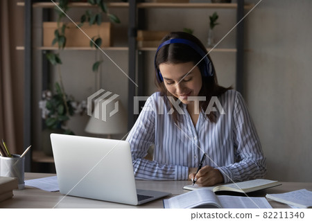 Smiling woman in headphones taking notes, studying online at home Smiling woman in headphones taking notes, studying online at home 82211340
